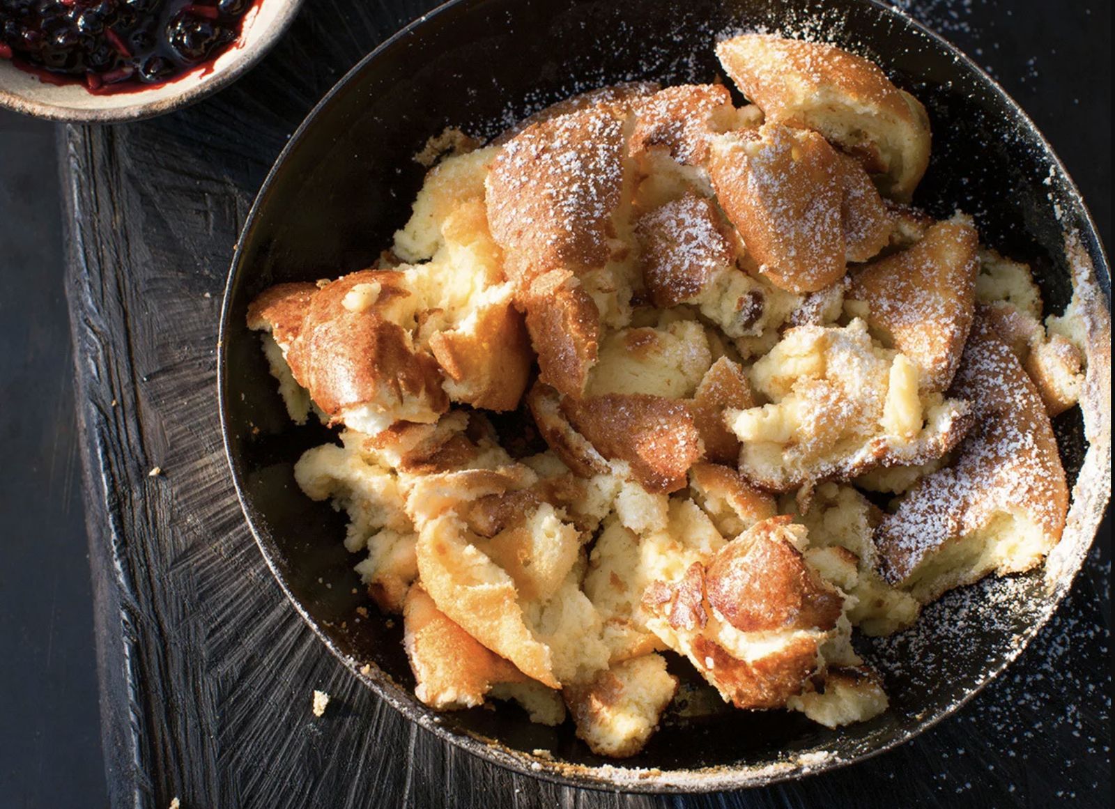 Kaiserschmarrn mit Puderzucker in einer schwarzen Eisenpfanne, daneben ein Schälchen dunkles Kompott